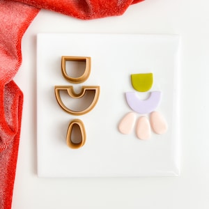 Może przedstawiać: Zestaw wykrawaczy do gliny polimerowej i kawałków gliny w różnych kształtach i kolorach. Wykrawacze są w odcieniach brązu, a kawałki gliny w kolorze zielonym, lawendowym i brzoskwiniowym. Ułożone na białej powierzchni z czerwonym aksamitnym materiałem w tle.