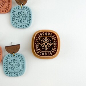 May include: Earrings with a square, floral design in shades of blue, brown, and tan. The earrings have a wooden top and a gold-colored hook. The design is repeated on a stamp.