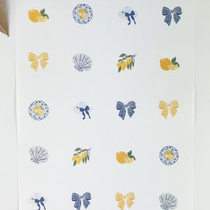 May include: A white sheet with repeating illustrations of lemons, bows, plates with lemons, and seashells. The illustrations are in yellow and blue, with a few green accents. The overall theme is a summery, coastal design.