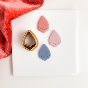 May include: Three polymer clay teardrop shapes in pink, blue, and coral on a white surface. A brown plastic cookie cutter in the shape of a teardrop is also on the surface.