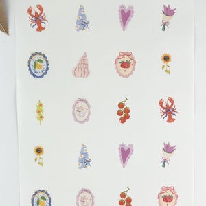 Peut inclure: Une feuille d'autocollants avec diverses illustrations. Les motifs comprennent un homard rouge, un gâteau bleu et blanc, un cœur violet, un bouquet de fleurs violettes, un citron, un coquillage, un emblème de fraise, un tournesol et une brochette d'olives.