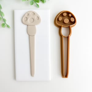 May include: A brown plastic mushroom cookie cutter and a beige silicone mushroom mold. Both are on a white surface.