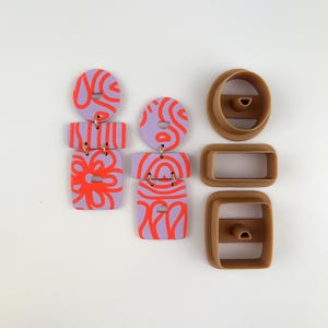 May include: Handmade polymer clay earrings in a modern design. The earrings feature a lavender base with red abstract patterns and gold-colored hardware. Also shown are brown clay cutters in oval and rectangular shapes.