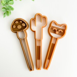 May include: Three brown plastic cookie cutters shaped like a mushroom, a cactus, and a raccoon.