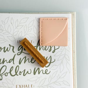 May include: A light-colored book with green script and leaf designs, featuring a small, square, peach-colored object with stitched edges and a rectangular, brown object with a ridged design. The text on the book reads "IN E: our goodness and me follow me EXHALE: all the days."