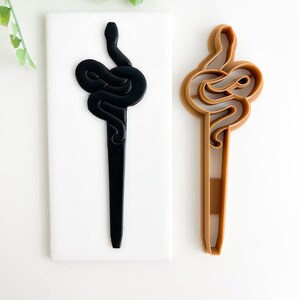 May include: A black and brown snake-shaped cookie cutter. The cutter is made of silicone and has a detailed design.