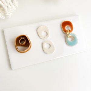 May include: A white ceramic tile with three ceramic shapes and a brown plastic mold. The ceramic shapes are two white ovals with a hole in the center and one blue and orange oval with a gold ring.