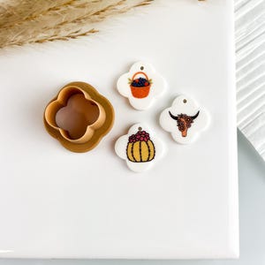 May include: A collection of craft supplies, including a brown four-leaf clover-shaped cookie cutter and three white, four-leaf clover-shaped charms. The charms feature illustrations of a basket of berries, a cactus, and a longhorn.