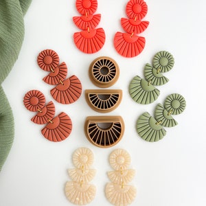 May include: A set of six pairs of handmade polymer clay earrings. The earrings are in three different colors: red, green, and cream. Each pair features a three-tiered design with a sunburst shape at the top and a half-circle shape at the bottom.