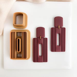Peut inclure: Deux paires de boucles d'oreilles en argile polymère de couleur bordeaux avec un design géométrique. Les boucles d'oreilles ont une forme rectangulaire avec une ouverture verticale au centre. Les boucles d'oreilles sont présentées à côté d'une paire de moules à boucles d'oreilles en argile polymère.
