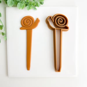 May include: Two brown plastic cookie cutters shaped like snails. The cutters are on a white surface.