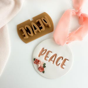 May include: A white ceramic ornament with the word "peace" in gold lettering and a cluster of pink and orange felt flowers. A brown silicone mold with the word "peace" is also shown.
