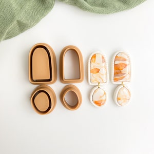 May include: A set of four brown silicone molds for making earrings, along with two pairs of finished earrings. The earrings are white with orange marbling and have a geometric shape.