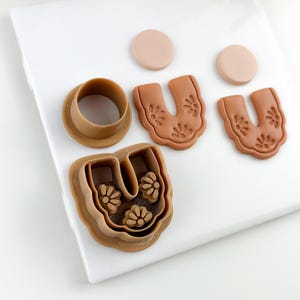May include: A collection of brown polymer clay cutters and shapes for crafting. Includes a round cutter, two U-shaped cutters with floral designs, two small circles, and a U-shaped cutter with floral details. The items are arranged on a white surface.