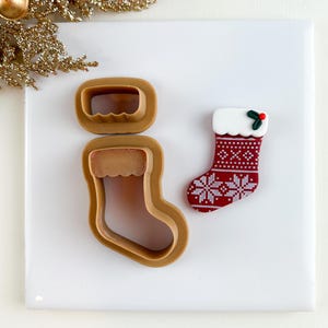 May include: A brown plastic cookie cutter set in the shape of a Christmas stocking, with a matching stocking-shaped cookie. The cookie is red with white snowflake patterns and a white cuff, topped with a sprig of holly.