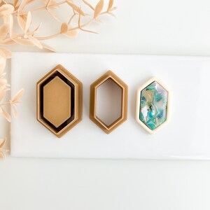 Puede incluir: Tres cortadores de galletas de forma hexagonal, dos son marrones y uno es blanco con un diseño marmoleado azul y verde.