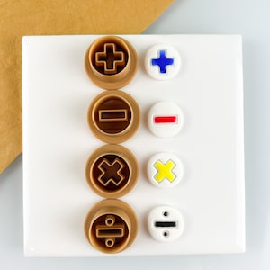 Könnte beinhalten: Eine Sammlung von vier braunen Ausstechern mit mathematischen Symbolen: Plus, Minus, Multiplikation und Division. Neben jedem Ausstecher befindet sich ein passender weißer Knopf mit demselben Symbol in Blau, Rot, Gelb und Schwarz. Auf einem weißen Quadrat angeordnet.