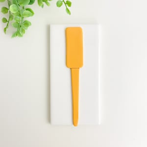 May include: A yellow ceramic utensil with a rounded rectangular shape and a long, thin handle. The utensil is sitting on a white surface.
