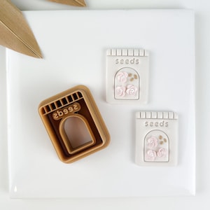 Puede incluir: Un cortador de galletas de madera con el número "26992" y dos decoraciones en forma de paquete de semillas. Los paquetes de semillas son blancos con detalles de flores rosas y la palabra "seeds" impresa. Los artículos están sobre una superficie blanca.