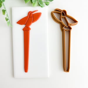 May include: An orange hummingbird cookie cutter next to a matching cookie. The hummingbird has outstretched wings and a long beak. The cookie cutter is made of brown plastic. The cookie is on a white surface.