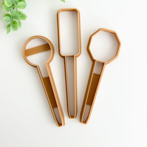 May include: Three brown plastic cookie cutters in the shape of a circle, rectangle, and octagon. The cutters are arranged in a fan shape on a white background.