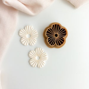 May include: Two white flower-shaped cookie cutters and a brown flower-shaped cookie cutter. The white cutters are made of plastic and the brown cutter is made of silicone.