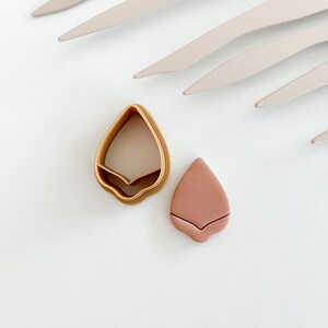 May include: A brown plastic cookie cutter shaped like a teardrop with a small indentation at the bottom, next to a pink clay teardrop shape.