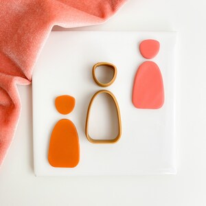 May include: Three polymer clay shapes, two orange and one pink, on a white surface. A gold metal cookie cutter in the shape of a person is also on the surface.