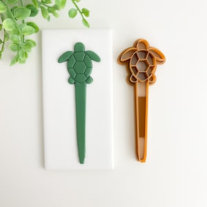 May include: Two green and brown plastic turtle cookie cutters. The cutters are on a white surface. The turtle on the left is solid green and the turtle on the right is hollow with a brown outline.