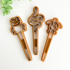 May include: Three brown plastic cookie cutters shaped like a frog, a snake, and a turtle.