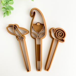 May include: Three brown plastic cookie cutters shaped like a gnome, a hummingbird, and a snail. The cutters are designed for baking and decorating cookies.