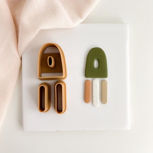May include: A set of polymer clay cutters and shapes in brown, green, white, and orange. The cutters are shaped like arches and rectangles. The shapes are a large arch, and three small rectangles.