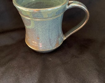 Puede incluir: Una taza de cerámica hecha a mano con un esmalte verde y marrón. La taza tiene una forma acampanada y una asa redondeada.