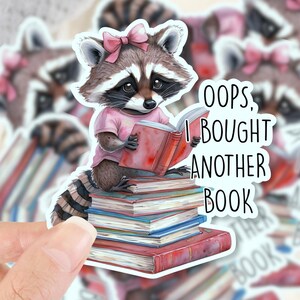 May include: A sticker featuring a raccoon wearing a pink shirt and bow, reading a book atop a stack of books. The text "Oops, I bought another book" is also on the sticker. Additional stickers are in the background.