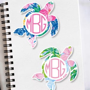 May include: Two turtle-shaped stickers with a tropical floral pattern in pink, blue, and green. Each sticker features a circular monogram with the letters "MBG" in pink. The stickers are on a white notebook.