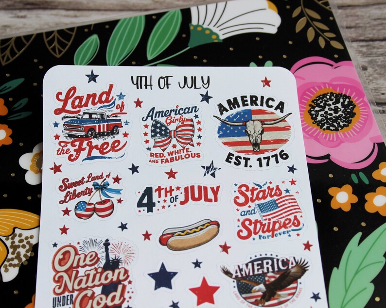 May include: A sheet of stickers with patriotic designs for the 4th of July. The stickers feature images of a truck, a bow, a steer skull, a hot dog, the American flag, and the Statue of Liberty. Text includes "Land of the Free", "America", and "Stars and Stripes".