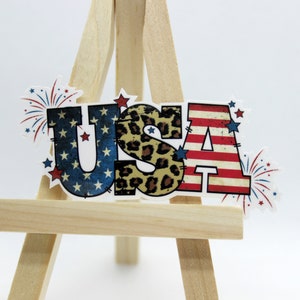 May include: A patriotic USA sign with red, white, and blue stars and stripes, a leopard print, and fireworks. The sign is on a small wooden easel.