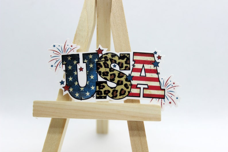 May include: A patriotic USA sign with red, white, and blue stars and stripes, a leopard print, and fireworks. The sign is on a small wooden easel.
