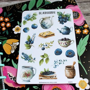 May include: A sticker sheet featuring watercolor illustrations of blueberries, teapots, a pie, and other blueberry-themed items. The word "BLUEBERRIES" is at the top. The sticker sheet is on a floral-patterned notebook.