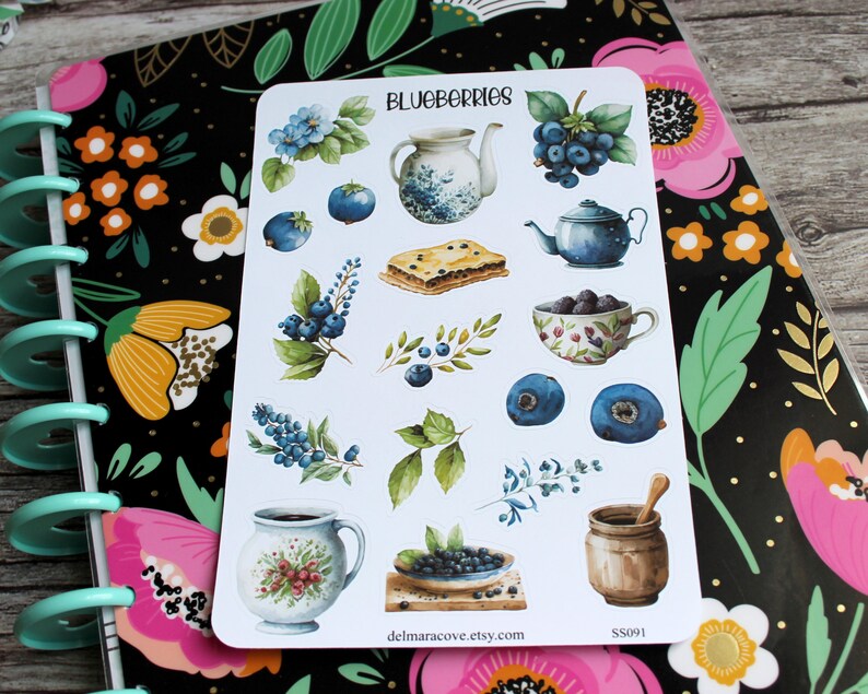 May include: A sticker sheet featuring watercolor illustrations of blueberries, teapots, a pie, and other blueberry-themed items. The word "BLUEBERRIES" is at the top. The sticker sheet is on a floral-patterned notebook.