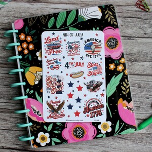 May include: A planner with a black floral cover and a sticker sheet featuring patriotic designs. The stickers include text like "4th of July," "America," and "Stars and Stripes," along with images of a hot dog, eagle, and the Statue of Liberty.