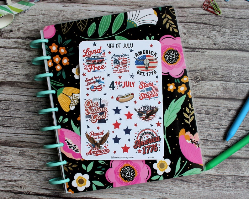 May include: A planner with a black floral cover and a sticker sheet featuring patriotic designs. The stickers include text like "4th of July," "America," and "Stars and Stripes," along with images of a hot dog, eagle, and the Statue of Liberty.