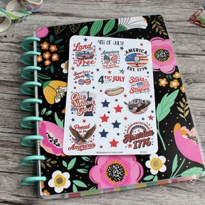 May include: A planner with a black floral cover, a teal spiral binding, and a sticker sheet featuring patriotic designs. The stickers include text like "4th of July," "America," and "Stars and Stripes," along with illustrations of a hot dog and an eagle.