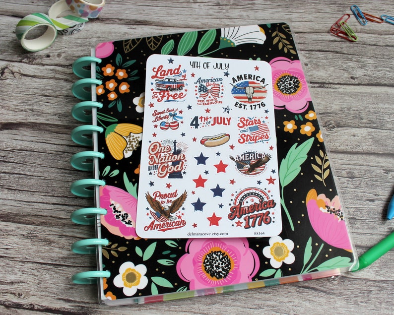 May include: A planner with a black floral cover, a teal spiral binding, and a sticker sheet featuring patriotic designs. The stickers include text like "4th of July," "America," and "Stars and Stripes," along with illustrations of a hot dog and an eagle.