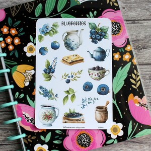 May include: A sticker sheet with watercolor illustrations of blueberries, teapots, and pastries. The text "BLUEBERRIES" is at the top. The sticker sheet is on a floral patterned notebook.