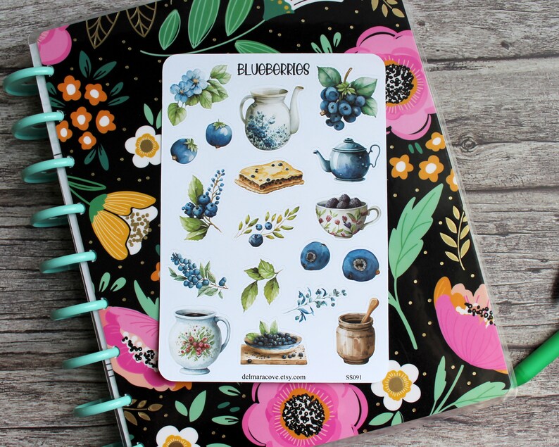 May include: A sticker sheet with watercolor illustrations of blueberries, teapots, and pastries. The text "BLUEBERRIES" is at the top. The sticker sheet is on a floral patterned notebook.