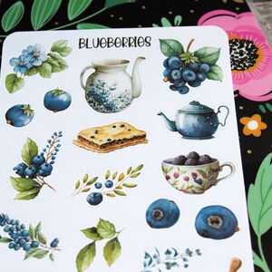 May include: A sheet of stickers featuring watercolor illustrations of blueberries, teapots, a slice of pie, and floral elements. The word "BLUEBERRIES" is written at the top. The stickers are on a white background.