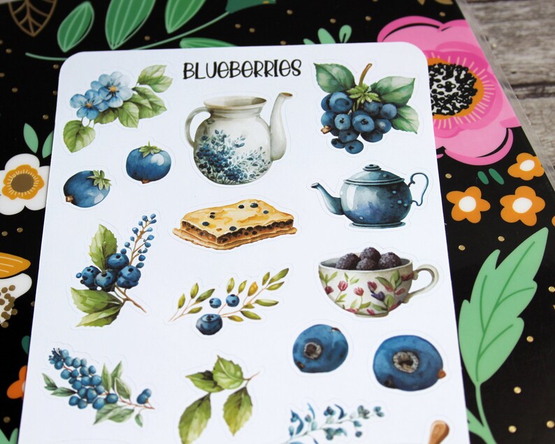 May include: A sheet of stickers featuring watercolor illustrations of blueberries, teapots, a slice of pie, and floral elements. The word "BLUEBERRIES" is written at the top. The stickers are on a white background.