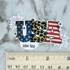 May include: A red, white, and blue USA sticker with a leopard print design in the center. The sticker has a pull here tab and is surrounded by red, white, and blue fireworks.
