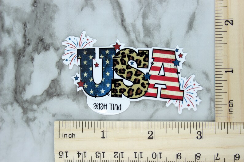 May include: A red, white, and blue USA sticker with a leopard print design in the center. The sticker has a pull here tab and is surrounded by red, white, and blue fireworks.